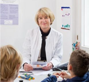 Prof. Dr. Anette-Gabriele Ziegler im weißen Kittel an ihrem Schreibtisch vor zwei Kindern sitzend