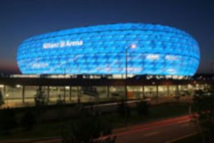 Nachtaufnahme der blauen Allianz Arena hinter Parkgarage und Autobahn mit roten Lichtstreifen.